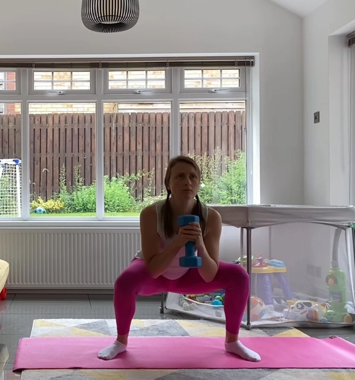 A woman performing a squat with a dumbbell on an exercise mat at home, with a baby playpen behind her